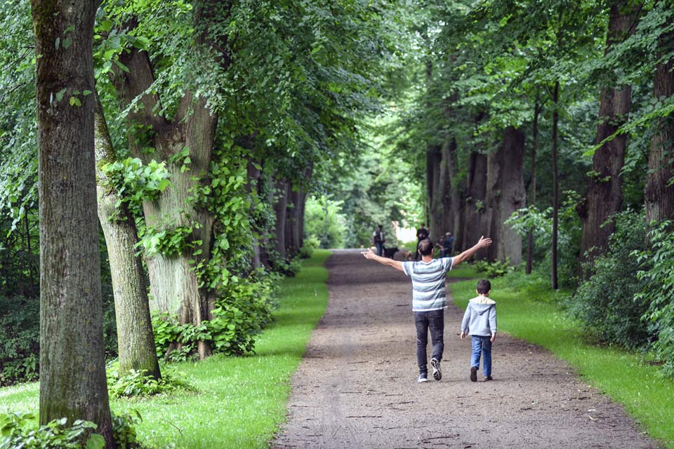 Ein Mann und ein Junge spazieren durch eine grün belaubte Allee, der Mann streckt beide Arme hoch in die Luft als wolle er die Welt oder wenigstens die herannahenden Spaziergänger mit ihren Hunden umarmen